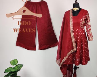 May include: A burgundy Indian salwar kameez set. The set includes wide-leg pants, a long-sleeved tunic with gold embroidery, and a matching dupatta. The tunic is displayed on a mannequin, and the pants are hanging on a hanger. The text "INDO WEAVES" is visible.