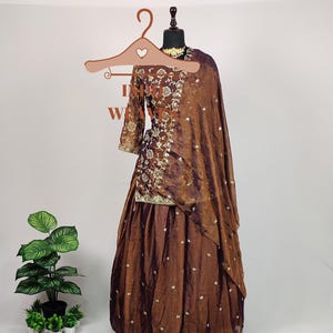 Può includere: Un completo formale ricamato marrone, composto da un top a maniche lunghe e una gonna fluida, esposto su un manichino. L'outfit presenta intricati ricami dorati e uno scialle abbinato. L'ensemble è progettato per occasioni speciali.