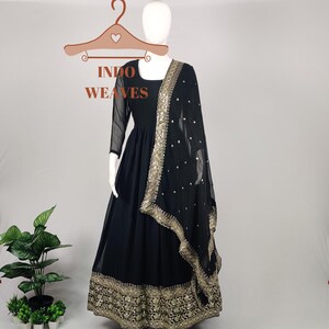 Puede incluir: Vestido negro largo hasta el suelo con bordados dorados y una bufanda transparente a juego. El vestido tiene un escote cuadrado y mangas largas. La bufanda está adornada con adornos dorados. El texto "INDO WEAVES" es visible.