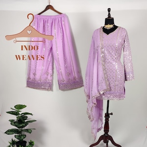 Puede incluir: Un conjunto lila claro de Indo Weaves, que incluye pantalones palazzo y una t&uacute;nica de manga larga con una bufanda a juego. El conjunto est&aacute; adornado con lentejuelas y ribetes dorados. Los pantalones palazzo est&aacute;n colgados en una percha de madera.