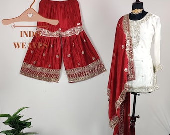 May include: A red and gold embroidered salwar kameez set. The set includes wide-legged trousers, a white tunic with silver embroidery, and a matching red scarf. The outfit is displayed on a mannequin and a hanger. The text "INDO WEAVES" is visible.
