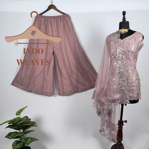 Può includere: Un completo pantalone palazzo color malva e una tunica abbinata impreziosita da una sciarpa trasparente sono esposti. La tunica è su un manichino, che mostra intricati dettagli di paillettes e pizzo. Il testo "INDO WEAVES" è visibile.