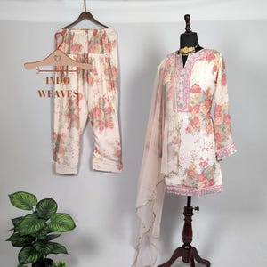 K&ouml;nnte beinhalten: Cremefarbenes Kurta-Anzug mit Blumenmuster, passender Hose und einem transparenten Schal. Das Outfit wird auf einer Schaufensterpuppe und einem Kleiderb&uuml;gel pr&auml;sentiert. Der Text "INDO WEAVES" ist auf dem Kleiderb&uuml;gel zu sehen. Das Blumenmuster umfasst rosa und rote Rosen.