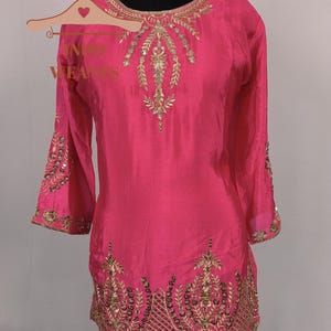Pu&ograve; includere: Una tunica rosa vibrante con ricami dorati. La tunica presenta intricati abbellimenti dorati intorno allo scollo, alle maniche e all'orlo. Il testo "INDO WEAVES" &egrave; visibile su un appendiabiti. &Egrave; presente anche una collana girocollo dorata.