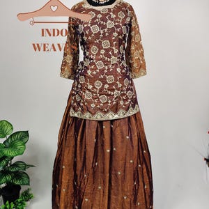 May include: A brown embroidered formal dress with a matching skirt. The dress features intricate floral patterns and gold accents. The sleeves are sheer, and the skirt has a flowing design. The text "INDO WEAV" is visible on a hanger.