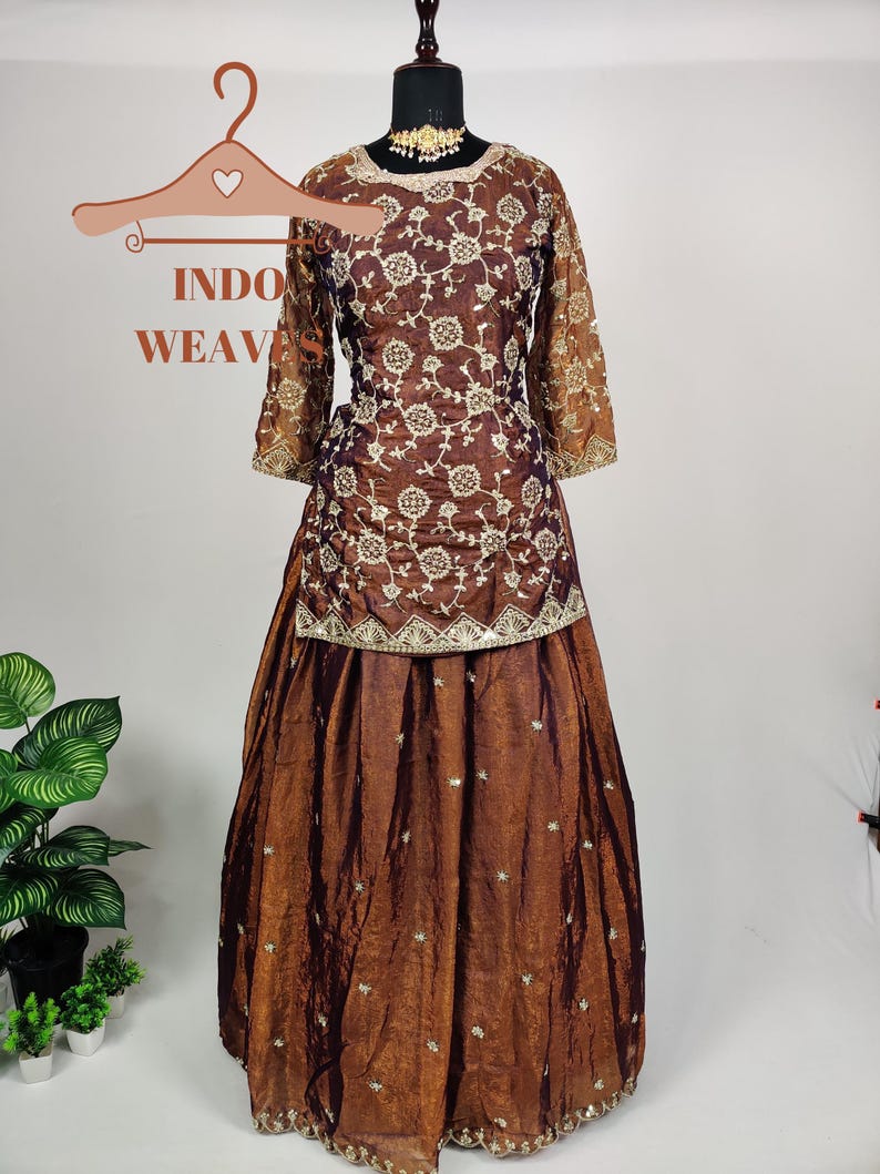 May include: A brown embroidered formal dress with a matching skirt. The dress features intricate floral patterns and gold accents. The sleeves are sheer, and the skirt has a flowing design. The text "INDO WEAV" is visible on a hanger.