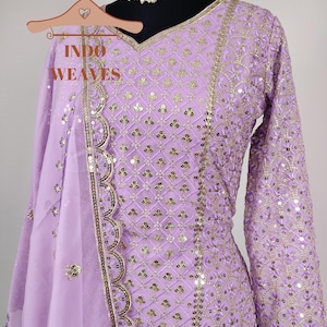Puede incluir: Un conjunto de salwar kameez de color lavanda con intrincados detalles de lentejuelas y bordados dorados. El atuendo presenta un escote en V, mangas largas y una dupatta a juego. El texto "INDO WEAVES" es visible en una percha.