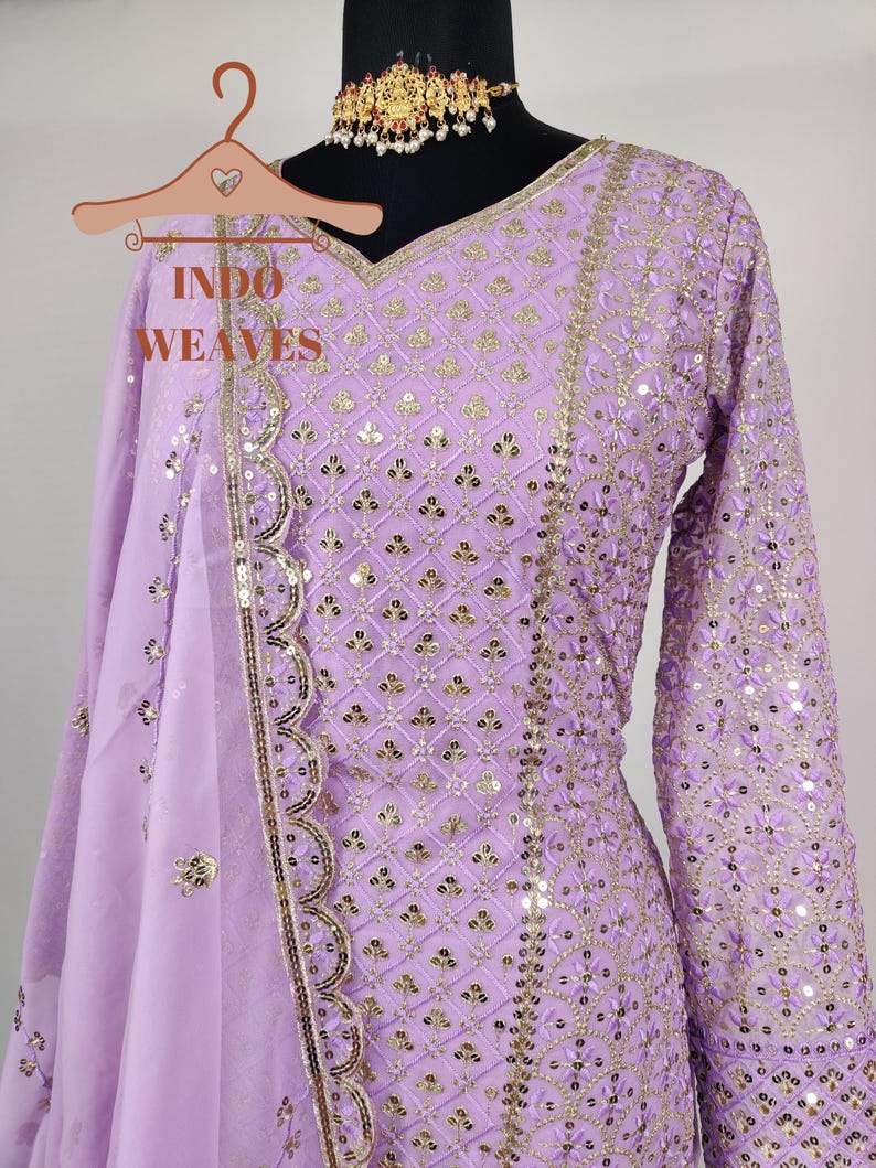 Puede incluir: Un conjunto de salwar kameez de color lavanda con intrincados detalles de lentejuelas y bordados dorados. El atuendo presenta un escote en V, mangas largas y una dupatta a juego. El texto "INDO WEAVES" es visible en una percha.
