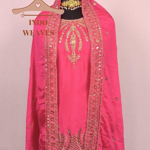 Pu&ograve; includere: Un vivace completo salwar kameez indiano rosa. L'outfit presenta intricati ricami dorati e dettagli di paillettes lungo i bordi del dupatta e sul corpetto. Il testo "INDO WEAVES" &egrave; visibile su un appendiabiti.