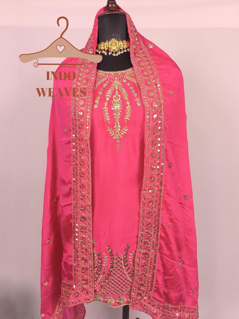 Pu&ograve; includere: Un vivace completo salwar kameez indiano rosa. L'outfit presenta intricati ricami dorati e dettagli di paillettes lungo i bordi del dupatta e sul corpetto. Il testo "INDO WEAVES" &egrave; visibile su un appendiabiti.