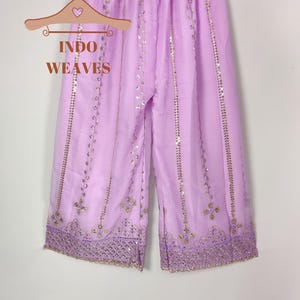 Puede incluir: Pantalones palazzo de color lila claro con detalles de lentejuelas y bordados dorados. Los pantalones est&aacute;n colgados de una percha de madera con el texto "INDO WEAVES". Los pantalones palazzo tienen un dise&ntilde;o de pierna ancha y est&aacute;n hechos de una tela ligera.