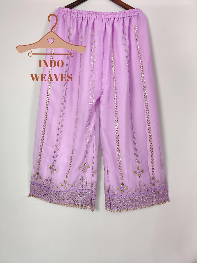 Puede incluir: Pantalones palazzo de color lila claro con detalles de lentejuelas y bordados dorados. Los pantalones est&aacute;n colgados de una percha de madera con el texto "INDO WEAVES". Los pantalones palazzo tienen un dise&ntilde;o de pierna ancha y est&aacute;n hechos de una tela ligera.