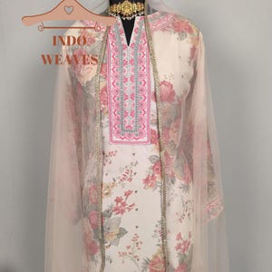 K&ouml;nnte beinhalten: Cremefarbener Salwar Kameez mit floralem Muster und rosa-silberner Stickerei am Ausschnitt und Besatz. Ein durchsichtiges, hellrosa Dupatta drapiert &uuml;ber den Schultern. Das Outfit wird auf einer Schaufensterpuppe pr&auml;sentiert.