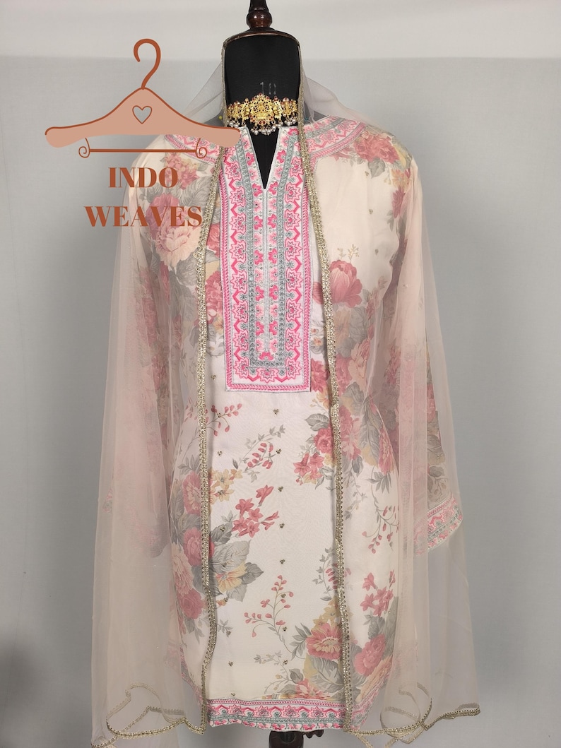 K&ouml;nnte beinhalten: Cremefarbener Salwar Kameez mit floralem Muster und rosa-silberner Stickerei am Ausschnitt und Besatz. Ein durchsichtiges, hellrosa Dupatta drapiert &uuml;ber den Schultern. Das Outfit wird auf einer Schaufensterpuppe pr&auml;sentiert.