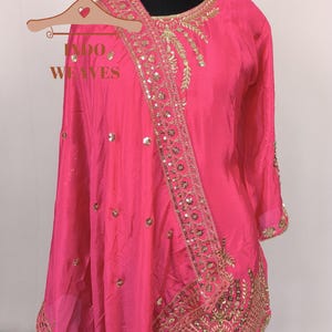 Pu&ograve; includere: Un vivace completo in seta rosa con ricami dorati e paillettes. L'ensemble include un top a maniche lunghe e una sciarpa abbinata, esposto su un manichino. Il testo "INDO WEAVES" &egrave; visibile su un appendiabiti in legno.