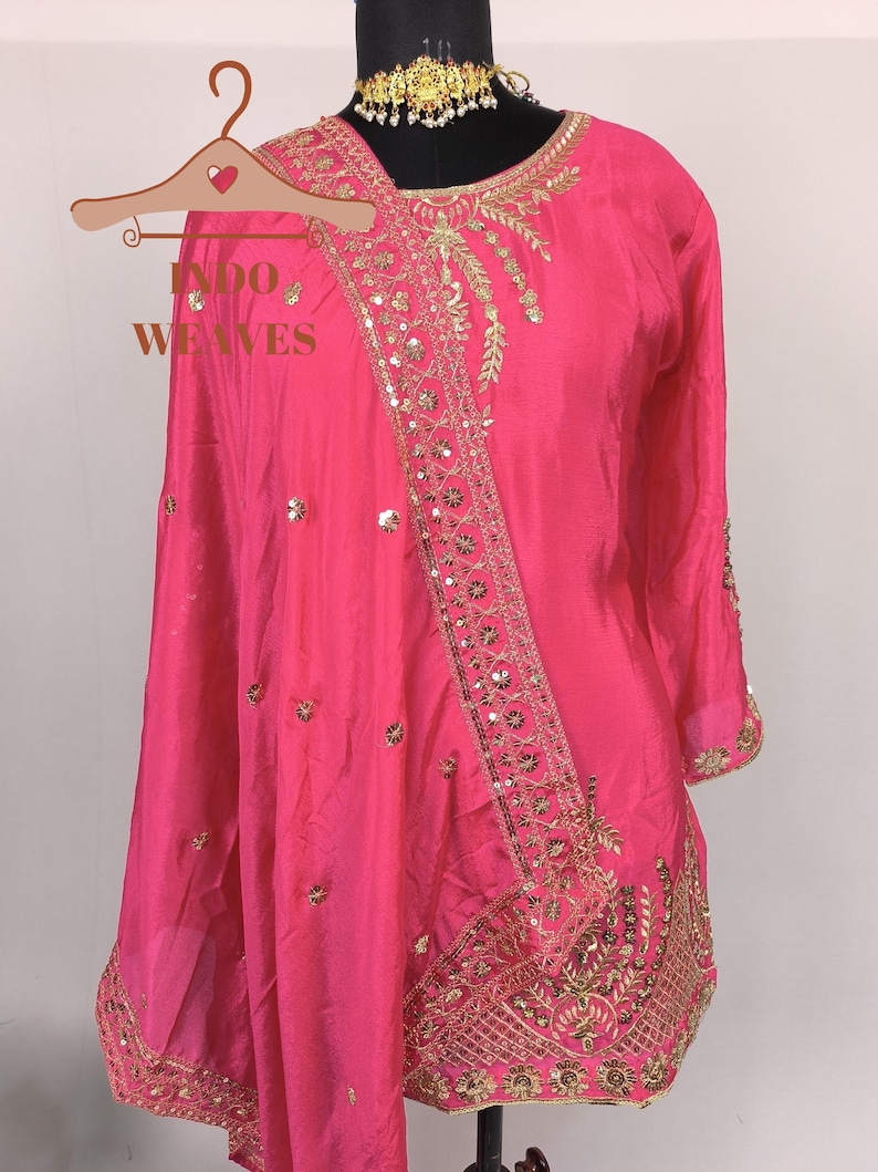 Pu&ograve; includere: Un vivace completo in seta rosa con ricami dorati e paillettes. L'ensemble include un top a maniche lunghe e una sciarpa abbinata, esposto su un manichino. Il testo "INDO WEAVES" &egrave; visibile su un appendiabiti in legno.