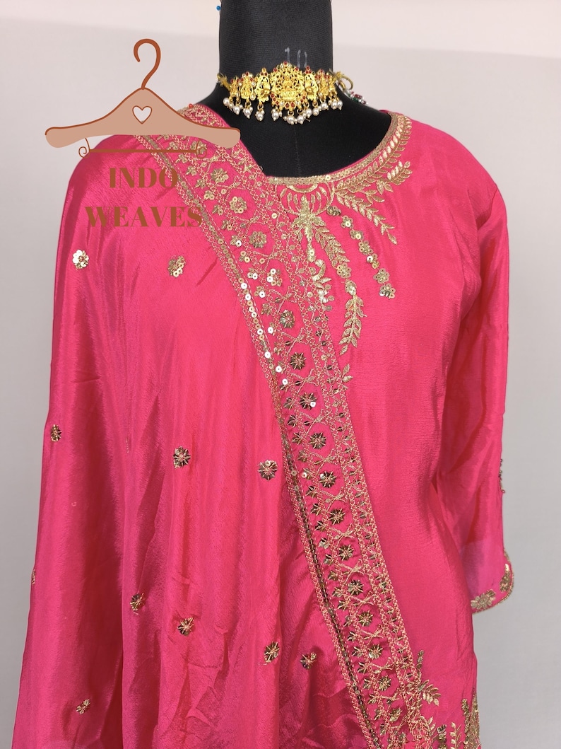 Pu&ograve; includere: Un vivace completo indiano rosa con intricati ricami dorati e abbellimenti di paillettes. Il completo presenta uno scollo tondo e maniche lunghe. &Egrave; visibile anche una collana girocollo dorata. Il testo "INDO WEAVES" &egrave; sull'appendiabiti.
