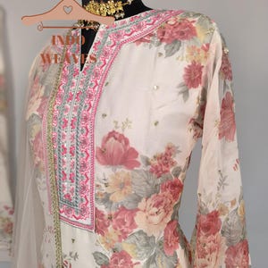 K&ouml;nnte beinhalten: Ein cremefarbener Anzug mit floralem Muster und V-Ausschnitt, verziert mit rosa und silberner Stickerei. Das lang&auml;rmlige Oberteil hat ein zartes Blumenmuster in Rosa-, Rot- und Gelbt&ouml;nen. Der Text "INDO WEAVES" ist auf dem Kleiderb&uuml;gel zu sehen.