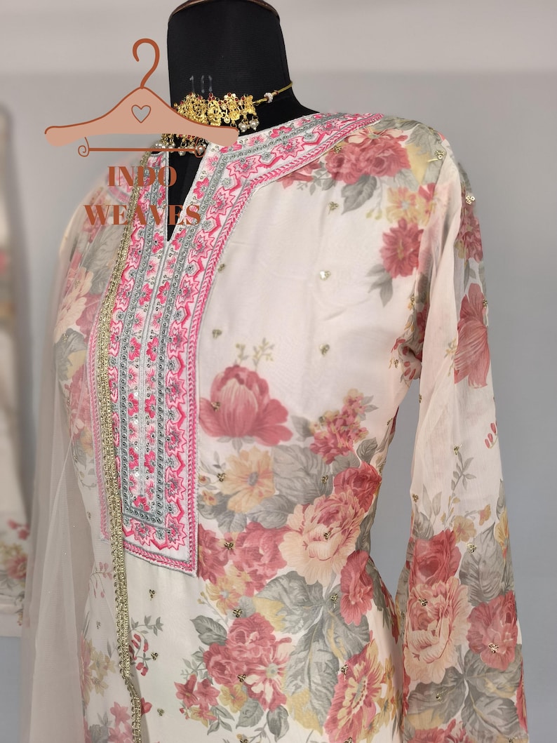 K&ouml;nnte beinhalten: Ein cremefarbener Anzug mit floralem Muster und V-Ausschnitt, verziert mit rosa und silberner Stickerei. Das lang&auml;rmlige Oberteil hat ein zartes Blumenmuster in Rosa-, Rot- und Gelbt&ouml;nen. Der Text "INDO WEAVES" ist auf dem Kleiderb&uuml;gel zu sehen.