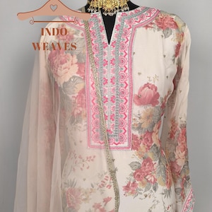 K&ouml;nnte beinhalten: Cremefarbenes Outfit mit floralem Muster und rosa und roten Blumen. Das Outfit hat einen V-Ausschnitt mit rosa und silberner Stickerei, lange &Auml;rmel und einen passenden, durchsichtigen Schal. Der Text "INDO WEAVES" ist auf einem Kleiderb&uuml;gel zu sehen.