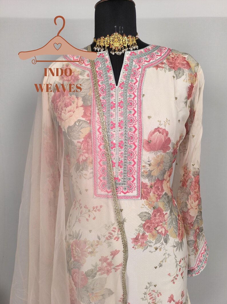 K&ouml;nnte beinhalten: Cremefarbenes Outfit mit floralem Muster und rosa und roten Blumen. Das Outfit hat einen V-Ausschnitt mit rosa und silberner Stickerei, lange &Auml;rmel und einen passenden, durchsichtigen Schal. Der Text "INDO WEAVES" ist auf einem Kleiderb&uuml;gel zu sehen.
