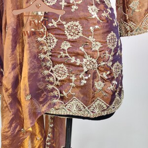 May include: A two-piece ensemble in shades of bronze and purple, featuring intricate gold embroidery. The top has a floral pattern and long sleeves. The matching scarf drapes elegantly. The fabric has a shimmering, iridescent quality.