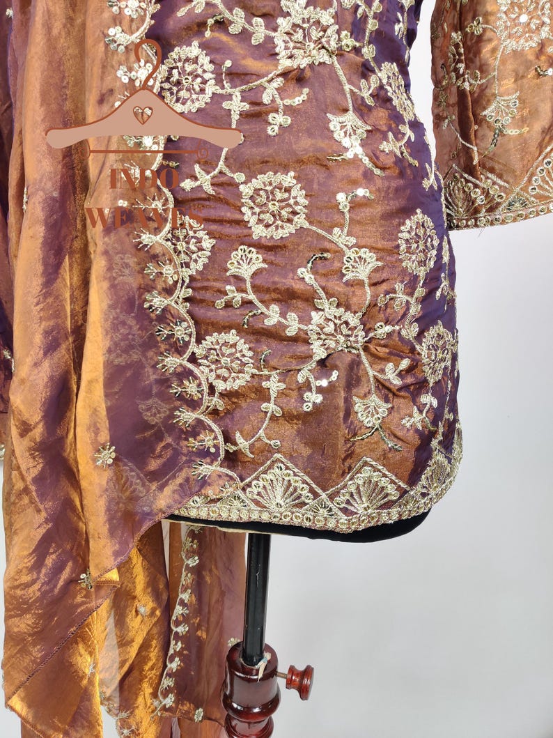 May include: A two-piece ensemble in shades of bronze and purple, featuring intricate gold embroidery. The top has a floral pattern and long sleeves. The matching scarf drapes elegantly. The fabric has a shimmering, iridescent quality.