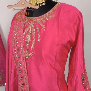 Pu&ograve; includere: Un vivace completo rosa a maniche lunghe con ricami dorati e paillettes. Lo scollo e le maniche presentano motivi dettagliati. Una collana dorata abbinata &egrave; esposta su una testa di manichino nera. L'outfit &egrave; esposto su una gruccia.