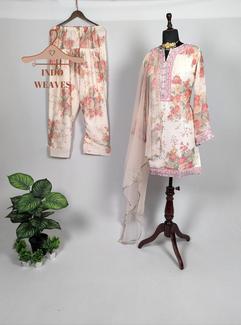 K&ouml;nnte beinhalten: Cremefarbenes Kurta-Anzug mit Blumenmuster, passender Hose und einem transparenten Schal. Das Outfit wird auf einer Schaufensterpuppe und einem Kleiderb&uuml;gel pr&auml;sentiert. Der Text "INDO WEAVES" ist auf dem Kleiderb&uuml;gel zu sehen. Das Blumenmuster umfasst rosa und rote Rosen.