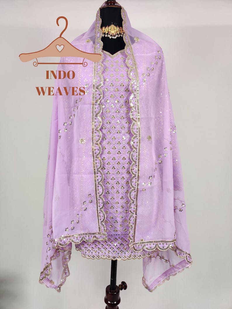 Puede incluir: Conjunto salwar kameez lila con detalles de lentejuelas y bordados dorados. El atuendo incluye una t&uacute;nica, una bufanda a juego y un collar gargantilla dorado. El texto "INDO WEAVES" es visible en una percha.