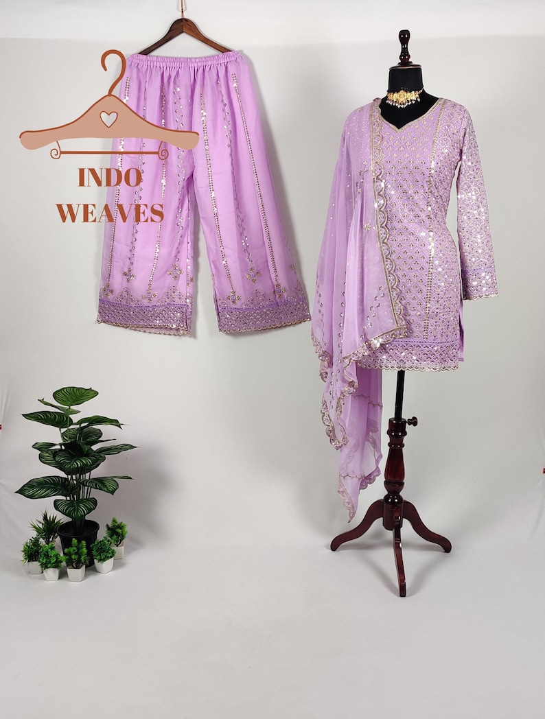 Puede incluir: Un conjunto lila claro de Indo Weaves, que incluye pantalones palazzo y una t&uacute;nica de manga larga con una bufanda a juego. El conjunto est&aacute; adornado con lentejuelas y ribetes dorados. Los pantalones palazzo est&aacute;n colgados en una percha de madera.