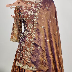 May include: A brown and purple embroidered formal dress with a matching shawl. The dress features intricate floral embroidery and a gold-colored necklace. The fabric has a shimmering effect, and the dress is displayed on a mannequin.
