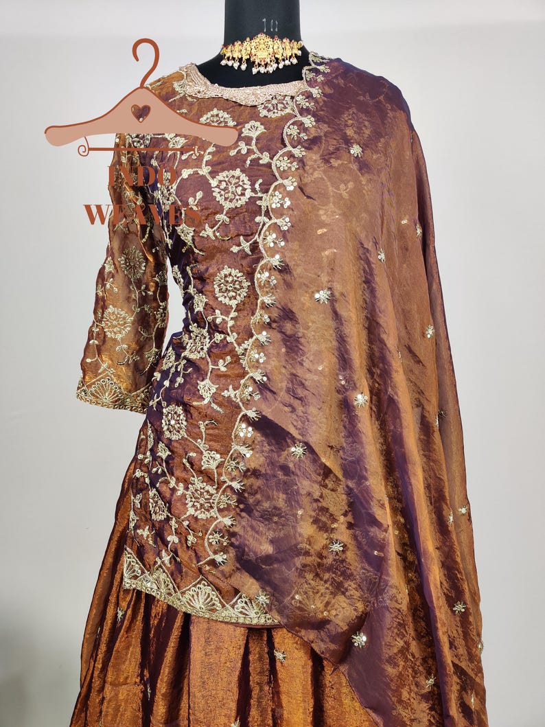 May include: A brown and purple embroidered formal dress with a matching shawl. The dress features intricate floral embroidery and a gold-colored necklace. The fabric has a shimmering effect, and the dress is displayed on a mannequin.