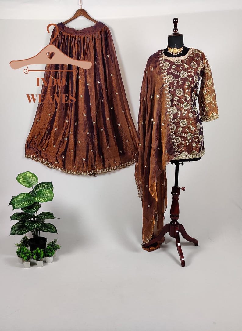 May include: A brown, two-piece ethnic outfit with intricate embroidery and embellishments. The ensemble includes a skirt and a tunic with a matching scarf. The outfit is displayed on a mannequin and a hanger, showcasing the details of the design.