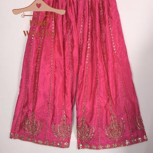 Pu&ograve; includere: Pantaloni palazzo rosa acceso con ricami dorati e paillettes. I pantaloni a gamba larga sono realizzati in un tessuto fluido e sono esposti su una gruccia di legno. I pantaloni presentano un design ornato sull'orlo.