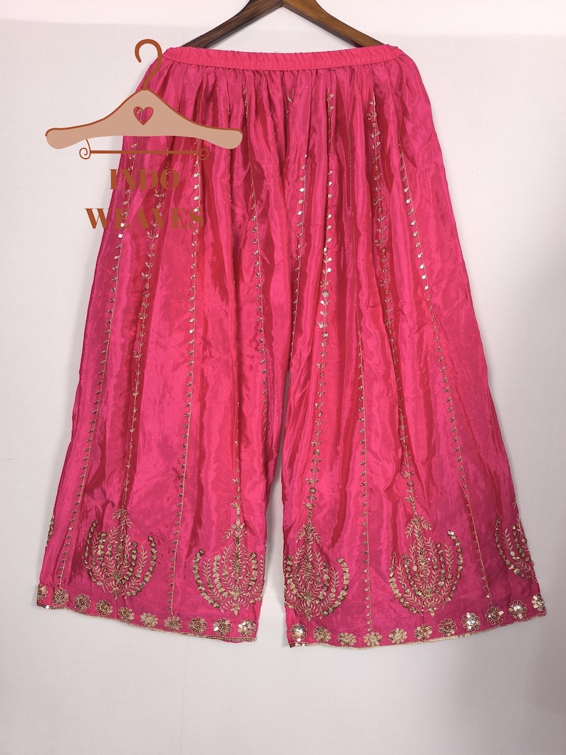 Pu&ograve; includere: Pantaloni palazzo rosa acceso con ricami dorati e paillettes. I pantaloni a gamba larga sono realizzati in un tessuto fluido e sono esposti su una gruccia di legno. I pantaloni presentano un design ornato sull'orlo.