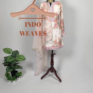 K&ouml;nnte beinhalten: Eine cremefarbene Tunika mit Blumendruck und ein passender, durchsichtiger Schal werden auf einer Schaufensterpuppe pr&auml;sentiert. Die Tunika hat lange &Auml;rmel und rosa und rote Blumenmuster. Der Text "INDO WEAVES" wird auf einem Kleiderb&uuml;gel angezeigt.