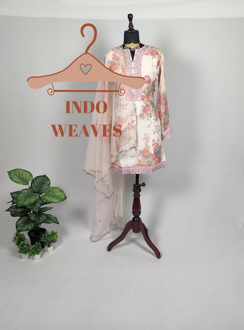 K&ouml;nnte beinhalten: Eine cremefarbene Tunika mit Blumendruck und ein passender, durchsichtiger Schal werden auf einer Schaufensterpuppe pr&auml;sentiert. Die Tunika hat lange &Auml;rmel und rosa und rote Blumenmuster. Der Text "INDO WEAVES" wird auf einem Kleiderb&uuml;gel angezeigt.