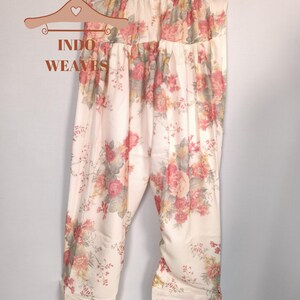K&ouml;nnte beinhalten: Cremefarbene Hose mit floralem Muster, die an einem Holzb&uuml;gel h&auml;ngt. Die Hose hat ein sich wiederholendes Muster aus rosa und roten Rosen mit gr&uuml;nen Bl&auml;ttern. Der Text "INDO WEAVES" befindet sich auf einem dekorativen Kleiderb&uuml;gel. Die Hose ist aus leichtem Stoff.