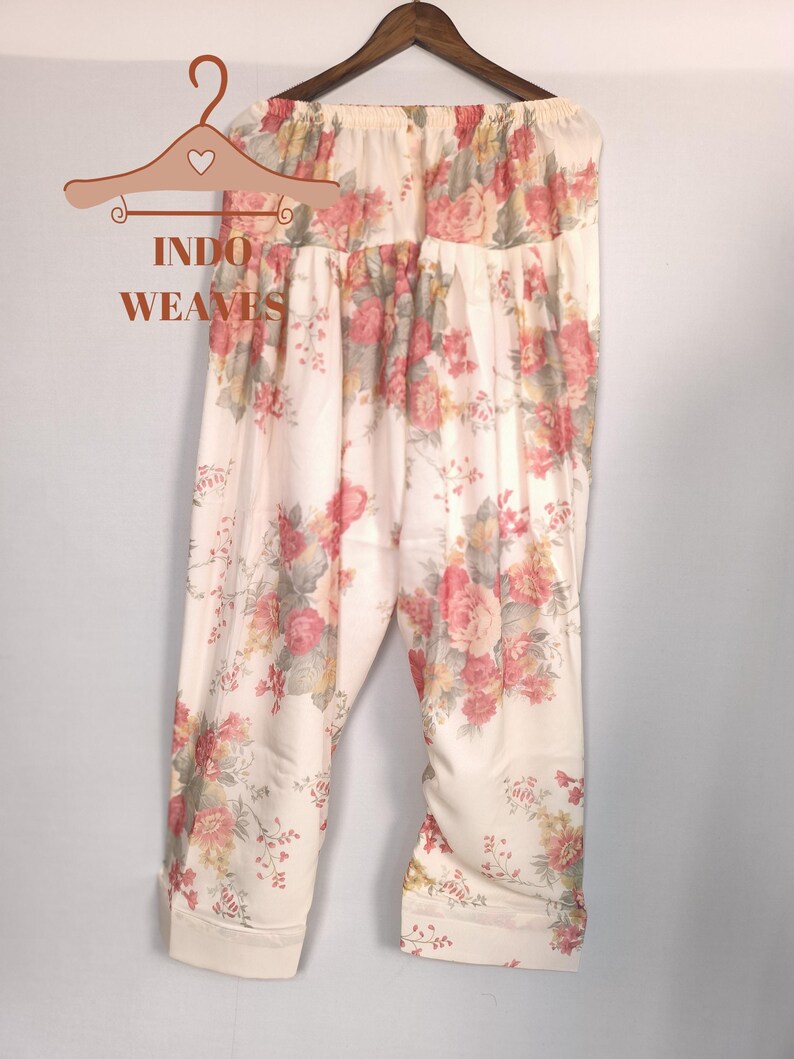 K&ouml;nnte beinhalten: Cremefarbene Hose mit floralem Muster, die an einem Holzb&uuml;gel h&auml;ngt. Die Hose hat ein sich wiederholendes Muster aus rosa und roten Rosen mit gr&uuml;nen Bl&auml;ttern. Der Text "INDO WEAVES" befindet sich auf einem dekorativen Kleiderb&uuml;gel. Die Hose ist aus leichtem Stoff.