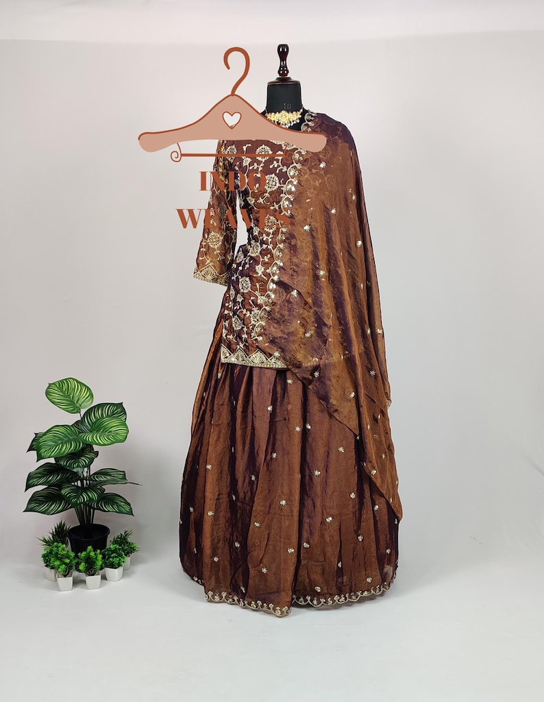 May include: A brown embroidered formal outfit, including a long-sleeved top and a flowing skirt, displayed on a mannequin. The outfit features intricate gold embroidery and a matching shawl. The ensemble is designed for special occasions.