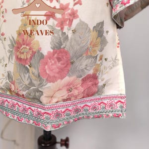 K&ouml;nnte beinhalten: Cremefarbenes Kleidungsst&uuml;ck mit floralem Druck mit rosa, gelben und grauen Blumen. Das Kleidungsst&uuml;ck hat einen dekorativen rosa und silbernen bestickten Saum und &Auml;rmelb&uuml;ndchen. Der Text "INDO WEAVES" ist sichtbar.