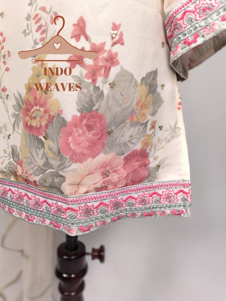 K&ouml;nnte beinhalten: Cremefarbenes Kleidungsst&uuml;ck mit floralem Druck mit rosa, gelben und grauen Blumen. Das Kleidungsst&uuml;ck hat einen dekorativen rosa und silbernen bestickten Saum und &Auml;rmelb&uuml;ndchen. Der Text "INDO WEAVES" ist sichtbar.