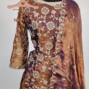 May include: A formal, two-piece outfit in shades of bronze and purple, featuring intricate floral embroidery. The ensemble includes a long-sleeved top and a draped skirt, with a matching shawl. A gold-toned necklace adorns the mannequin's neck.