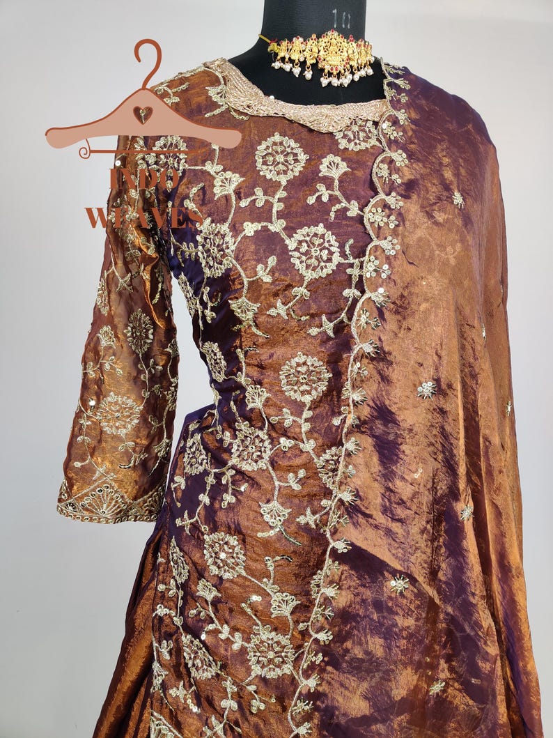 May include: A formal, two-piece outfit in shades of bronze and purple, featuring intricate floral embroidery. The ensemble includes a long-sleeved top and a draped skirt, with a matching shawl. A gold-toned necklace adorns the mannequin's neck.