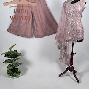 Può includere: Una tunica senza maniche color rosa polvere con ricami di paillettes argentate e un dupatta trasparente abbinato, accostata a pantaloni a gamba larga su una gruccia. Il set presenta motivi floreali e una collana dorata sul manichino.