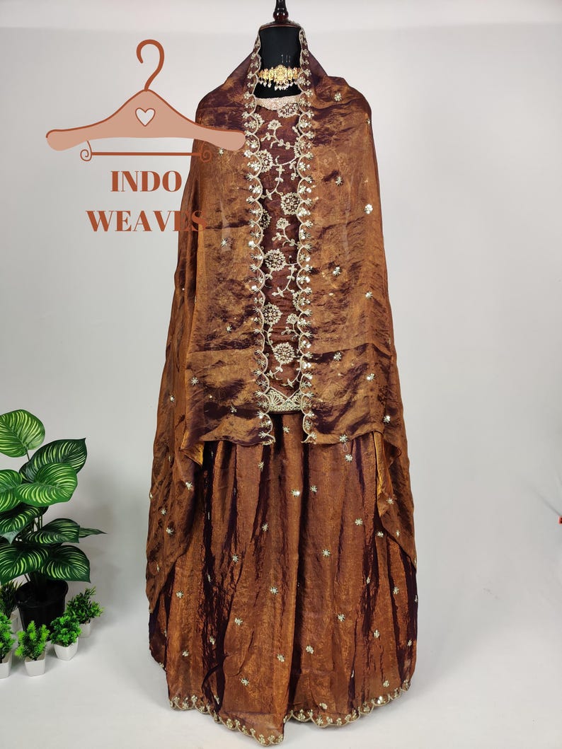 May include: A brown, formal dress with a matching shawl, displayed on a mannequin. The dress features intricate embroidery and embellishments. The text "INDO WEAVES" is visible on a hanger in the image.