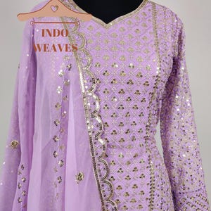 Puede incluir: Un conjunto de salwar kameez de color lavanda con intrincados detalles de lentejuelas y bordados dorados. El atuendo incluye una t&uacute;nica de manga larga, pantalones a juego y una dupatta transparente. El escote est&aacute; adornado con ribetes dorados. El texto "INDO WEAVES" es visible.