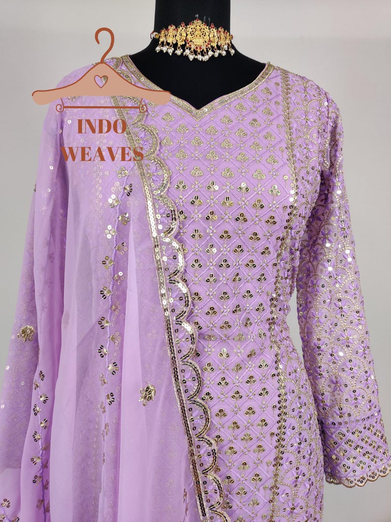 Puede incluir: Un conjunto de salwar kameez de color lavanda con intrincados detalles de lentejuelas y bordados dorados. El atuendo incluye una t&uacute;nica de manga larga, pantalones a juego y una dupatta transparente. El escote est&aacute; adornado con ribetes dorados. El texto "INDO WEAVES" es visible.
