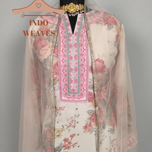 K&ouml;nnte beinhalten: Ein Salwar Kameez mit floralem Muster und passendem Dupatta. Der cremefarbene Stoff weist ein gro&szlig;es Muster aus rosa und roten Rosen auf. Der Ausschnitt und die Kanten sind mit Stickereien und goldenen Zierleisten verziert. Das Outfit wird auf einer Schaufenquin mit einer goldenen Halskette pr&auml;sentiert.